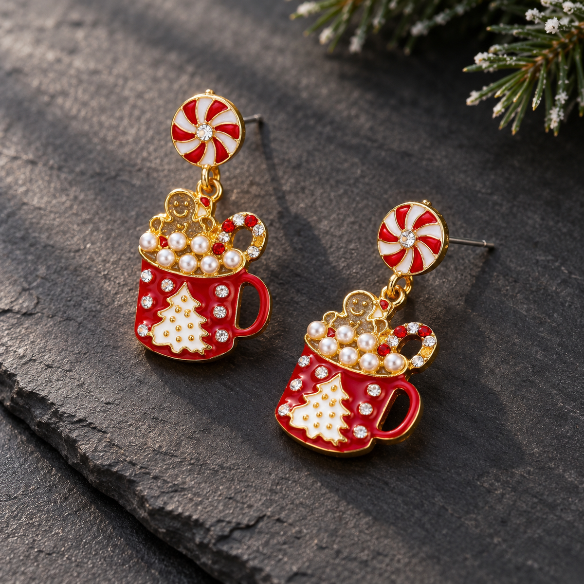 A pair of festive holiday dangle earrings featuring red and white peppermint candy studs, gold-tone chains, and red coffee mug charms decorated with white pearls, a white Christmas tree, and clear rhinestones, resting on a dark textured slate surface.