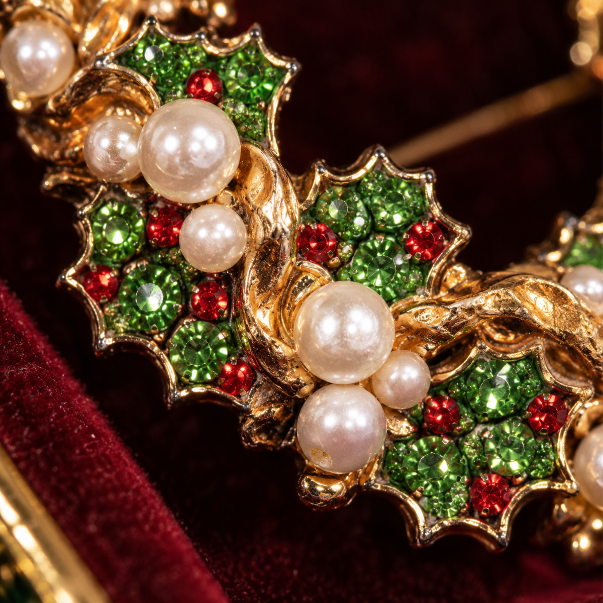 A close-up of a gold-toned Christmas holly necklace or brooch featuring green rhinestone-embellished holly leaves, white faux pearls, and small red rhinestones, displayed on a dark red velvet surface.