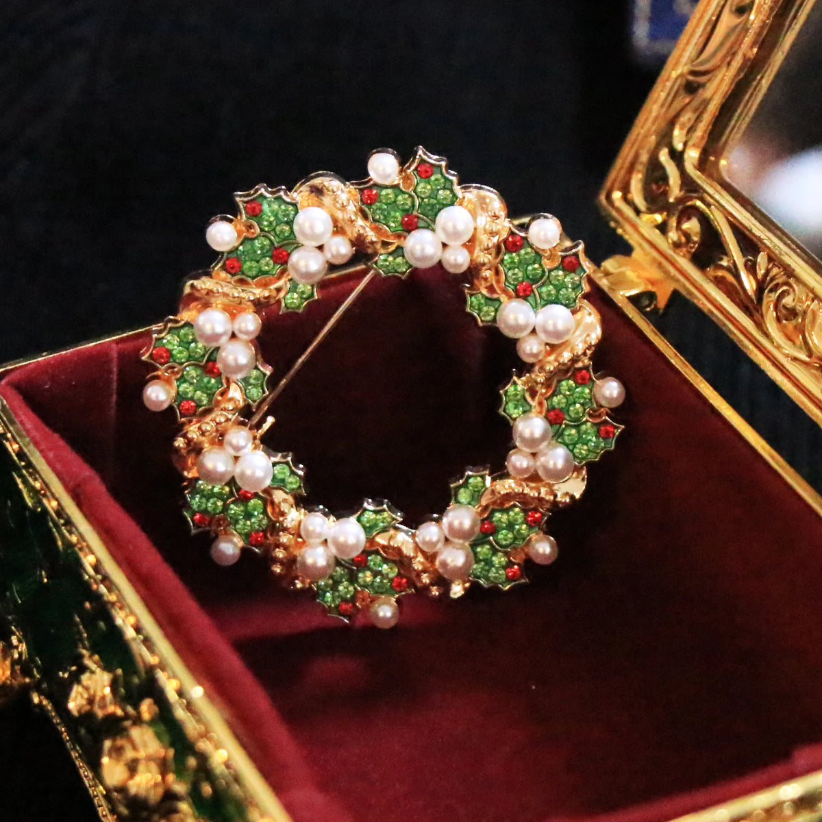 A gold-tone Christmas wreath brooch decorated with green enamel leaves, red enamel accents, and white faux pearls, displayed inside an ornate gold and red jewelry box.