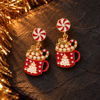 A pair of festive holiday dangle earrings featuring red and white peppermint candy swirls and cups decorated with gold-tone details, faux pearls, and white crystal embellishments, resting on a dark textured surface with warm golden tinsel in the background.