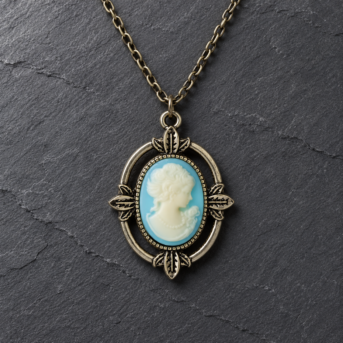 An antique bronze oval pendant necklace featuring a classic cameo-style portrait with a light blue and white background, displayed on a dark slate surface.