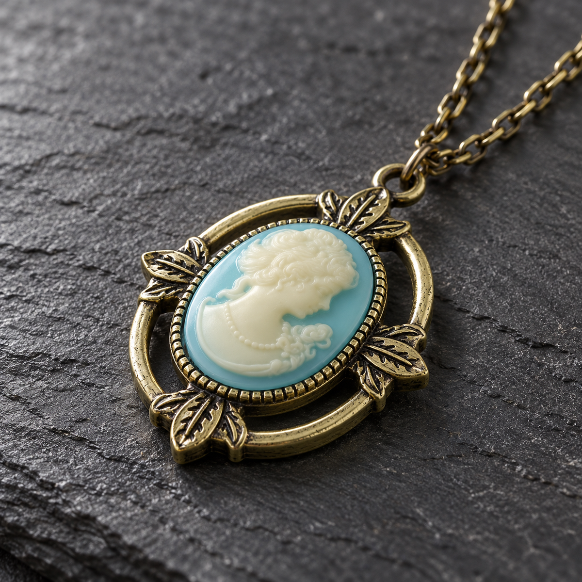 An oval pendant necklace with a light blue and cream cameo-style profile design, set in an ornate antique gold-tone metal frame with a matching chain, displayed on a dark textured slate surface.