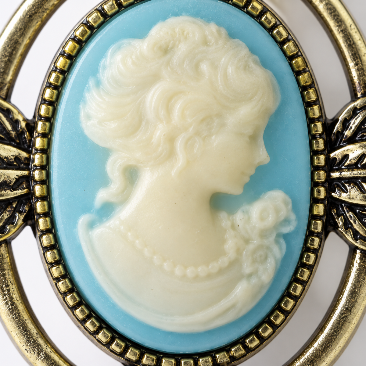 An antique-style oval cameo brooch featuring a cream-colored carved profile of a woman with a pearl necklace on a light blue background, surrounded by an ornate gold-toned metal frame.