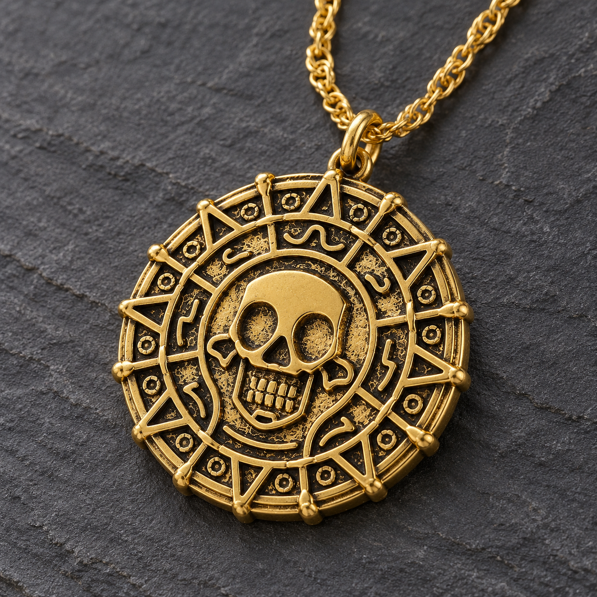 A gold-toned circular pendant necklace featuring a central skull motif and geometric border details, shown on a dark textured stone background.