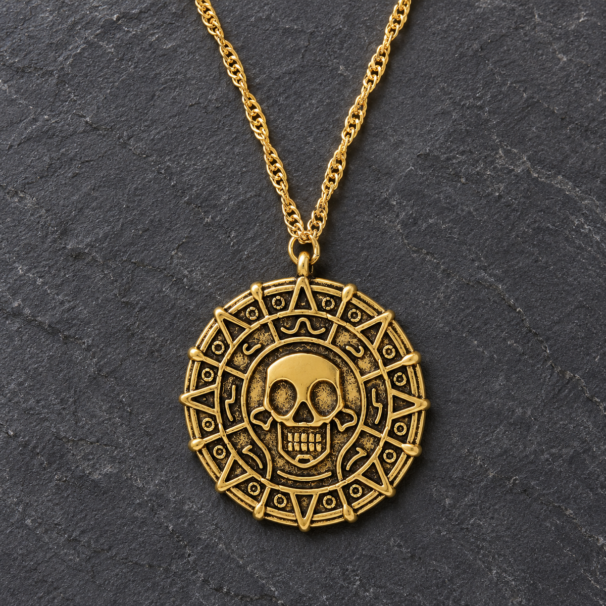 A gold-toned Aztec calendar pendant necklace with a skull in the center, displayed on a dark textured slate background.
