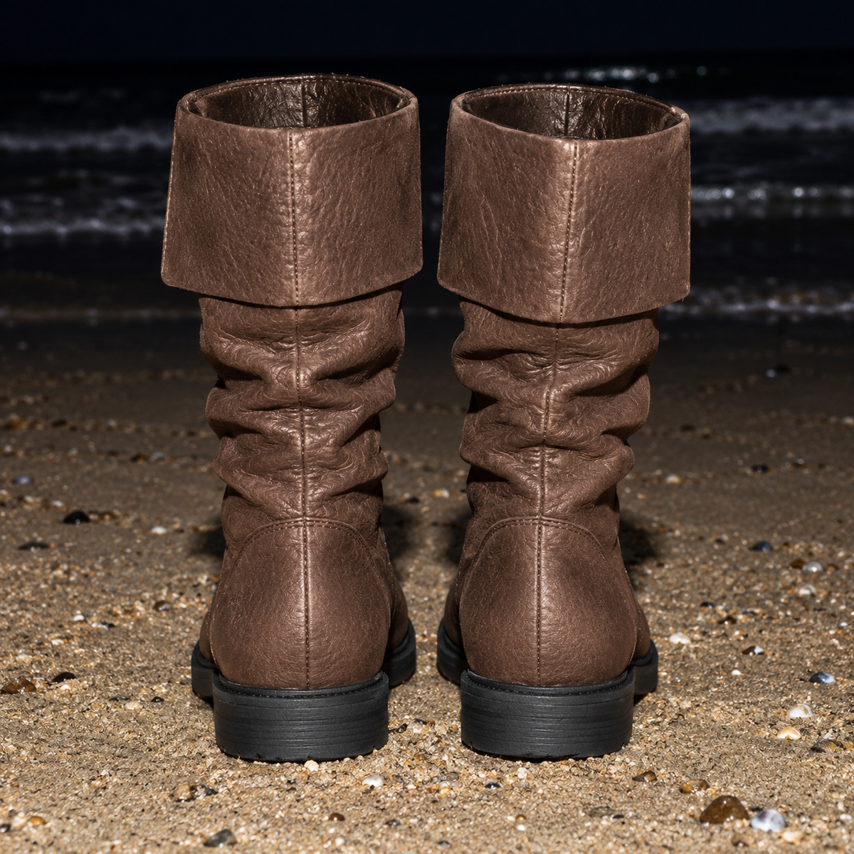 A pair of brown slouchy mid-calf boots with a wide folded cuff, shown from the back on a sandy beach with dark water in the background.