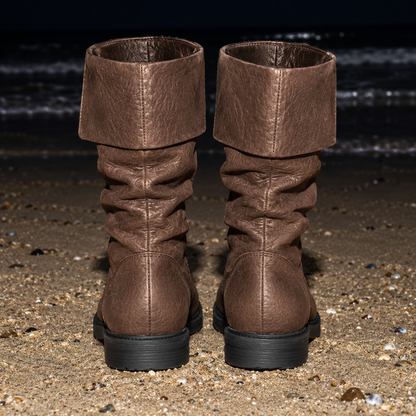 A pair of brown slouchy mid-calf boots with a wide folded cuff, shown from the back on a sandy beach with dark water in the background.