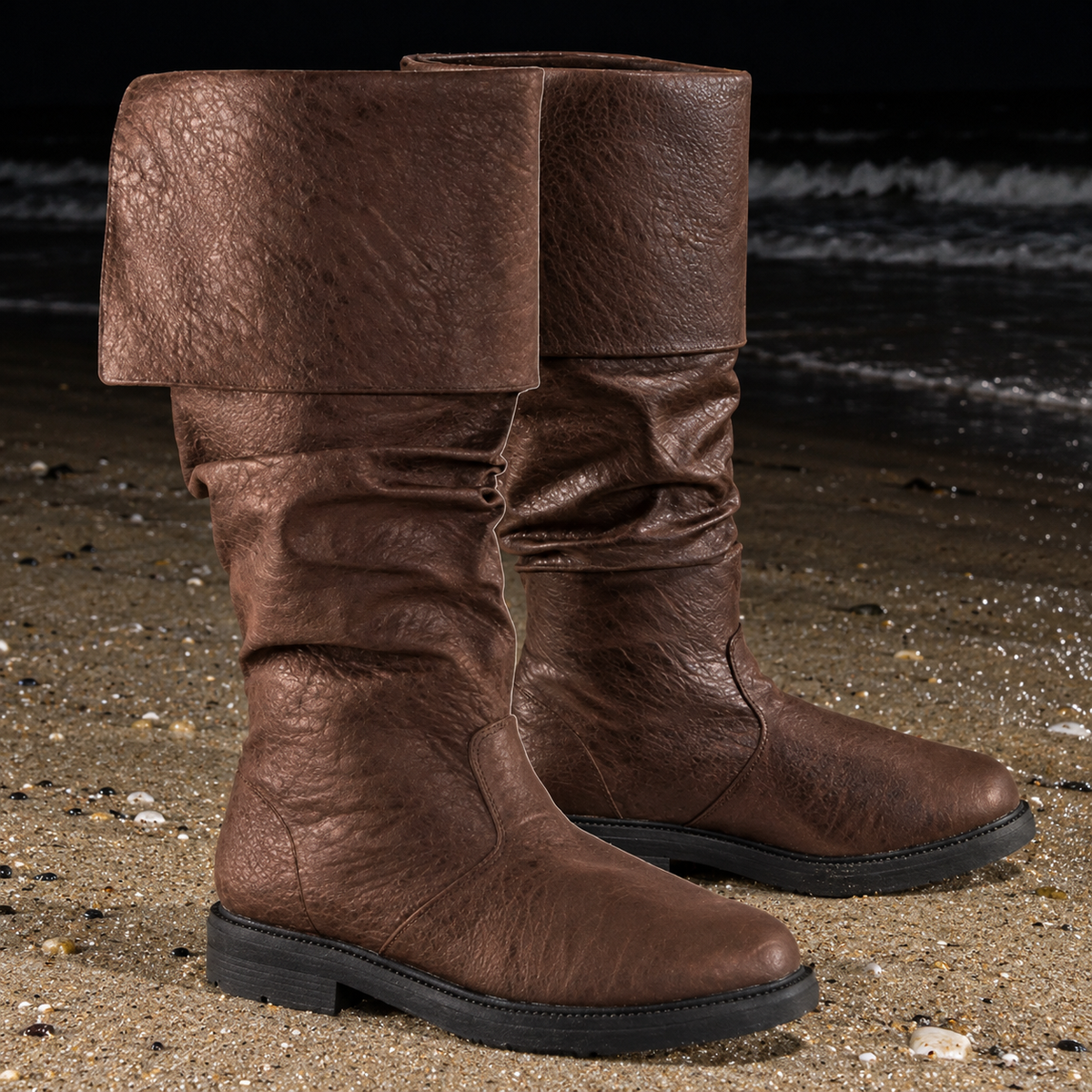 A pair of brown slouchy mid-calf boots with a wide folded cuff, shown on a sandy beach with the ocean in the background.