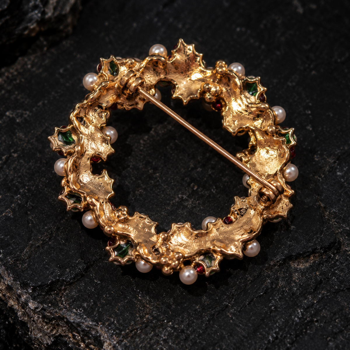 A circular gold holly leaf brooch decorated with small white pearls and green gemstones, displayed on a dark textured stone surface.