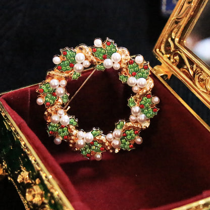 A gold-tone Christmas wreath brooch decorated with green enamel leaves, red enamel accents, and white faux pearls, displayed inside an ornate gold and red jewelry box.