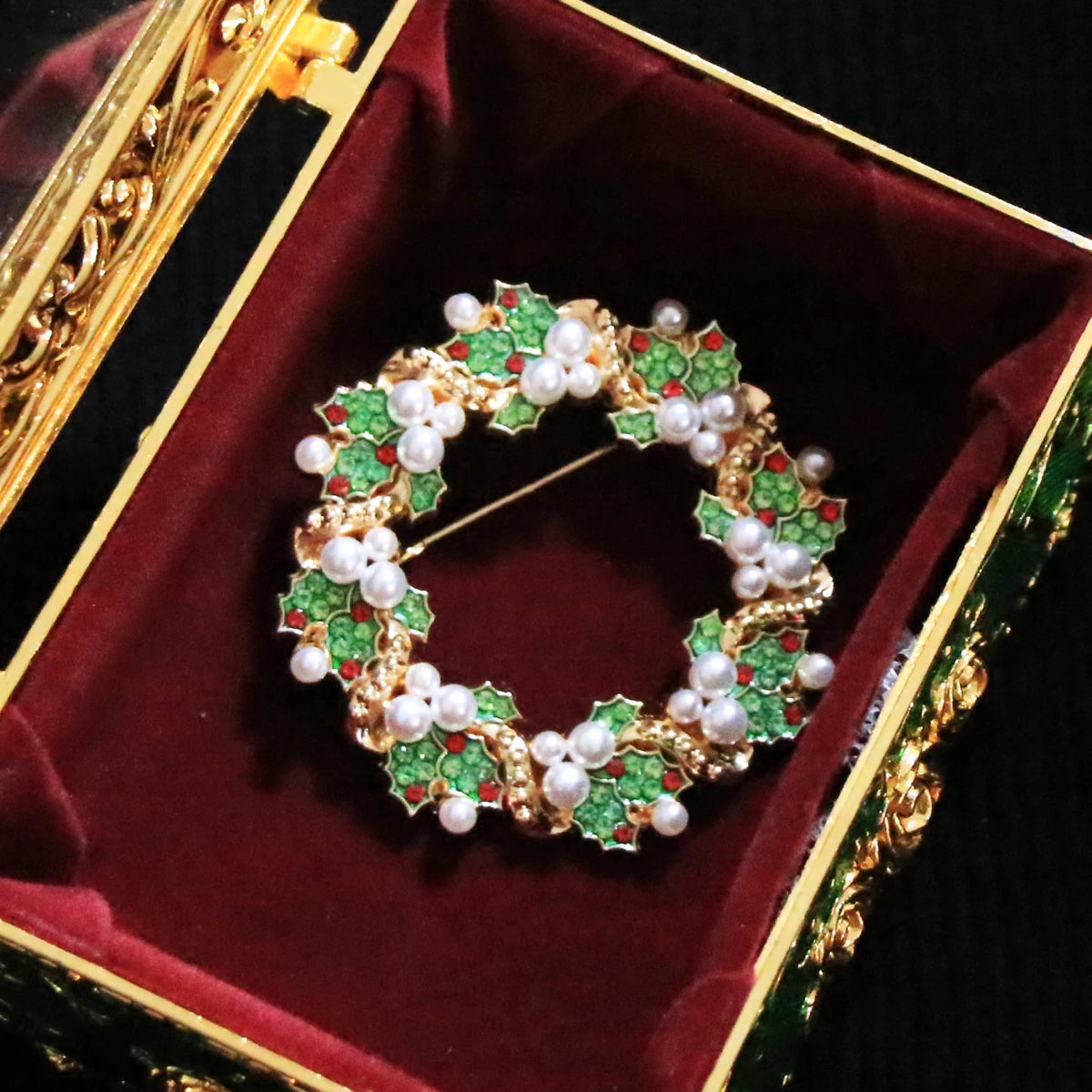 A gold holly wreath brooch decorated with green enamel, white pearls, and small red accents, displayed inside an ornate green and gold jewelry box with a burgundy velvet lining.