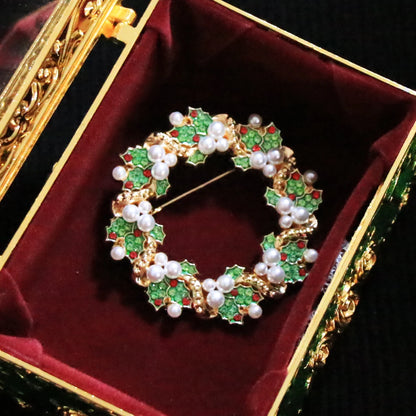 A gold holly wreath brooch decorated with green enamel, white pearls, and small red accents, displayed inside an ornate green and gold jewelry box with a burgundy velvet lining.