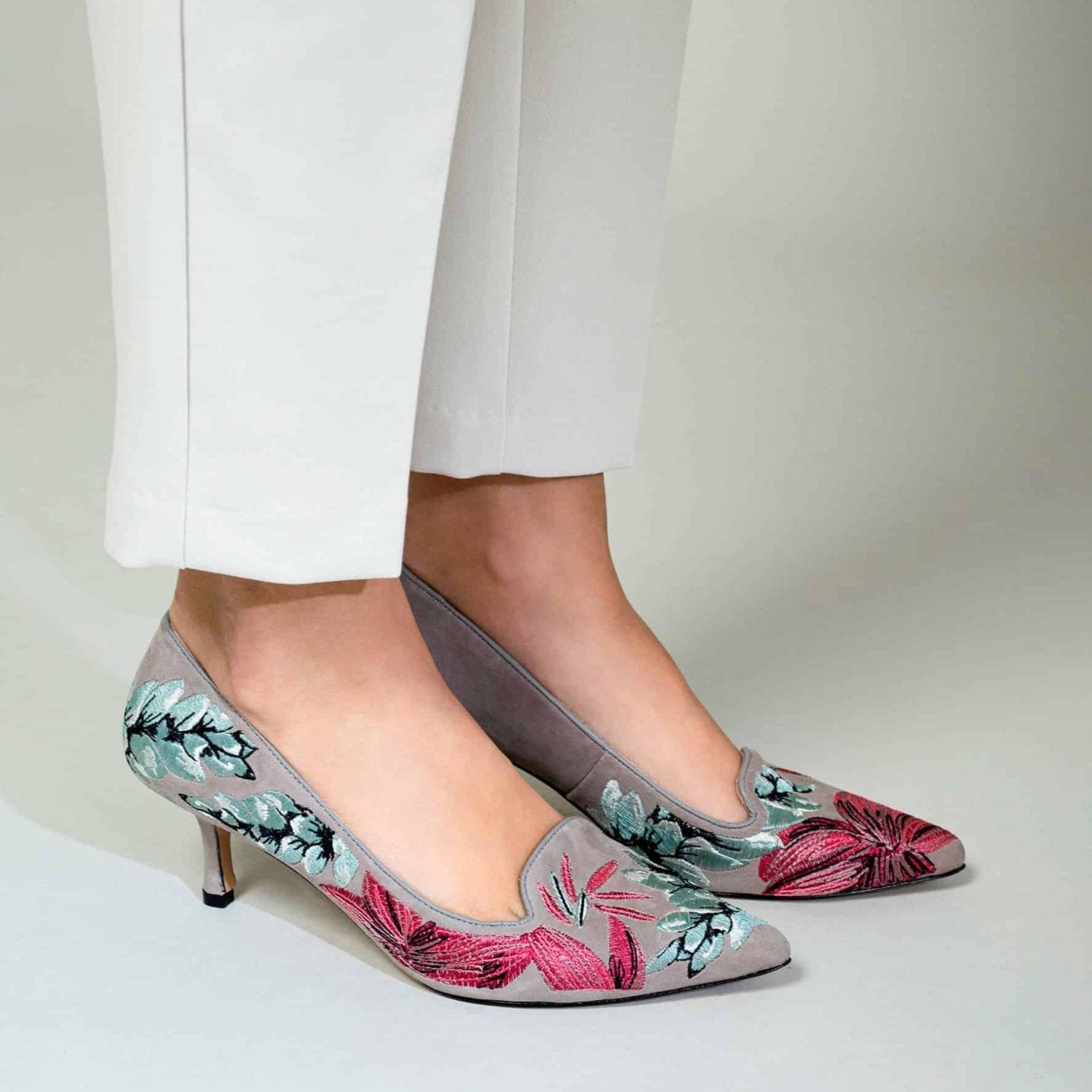 Gray high-heeled shoes with floral design worn by a woman in white pants on a light gray background