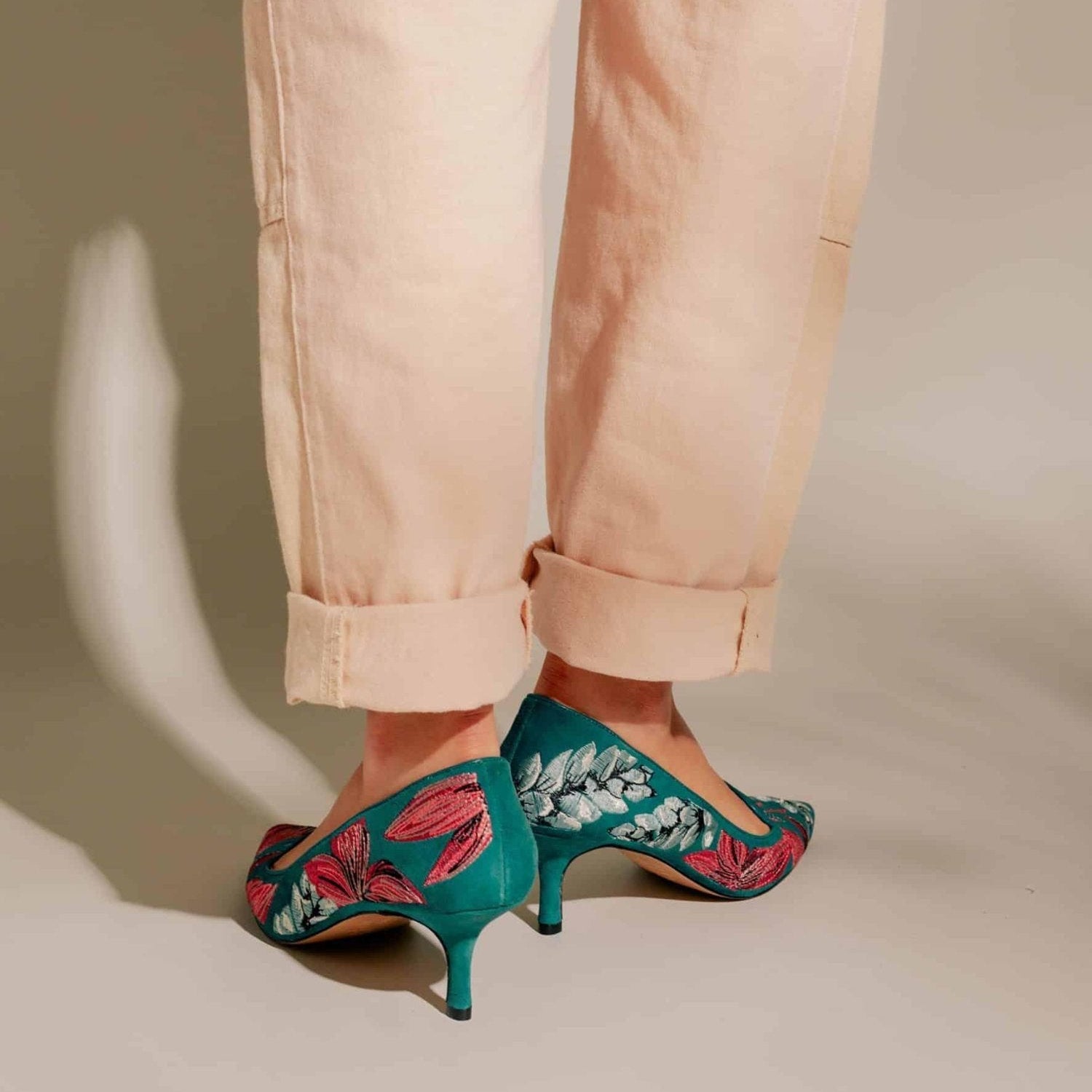 Teal high-heeled shoes with floral embroidery worn with beige pants on a neutral background